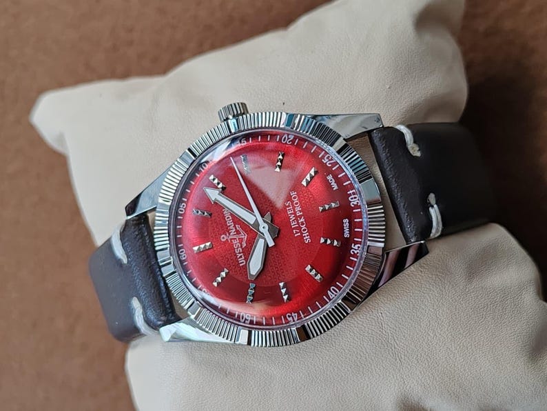 May include: A wristwatch with a vibrant red dial, silver-tone hands, and hour markers. The face features the text Ulysse Nardin, 17 Jewels, and Shock Proof. It has a silver fluted bezel and a dark brown leather strap with white stitching.