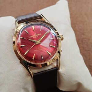 May include: A gold-tone wristwatch with a vibrant red dial featuring a textured pattern, gold-tone hands, and hour markers. The dial displays the text ULYSSE NARDIN, 17 JEWELS, SHOCK PROOF, and SWISS MADE. It includes a dark brown leather strap.