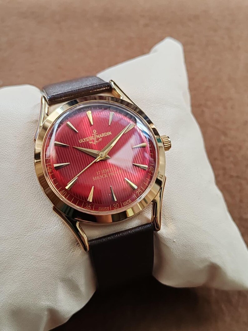 May include: A gold-tone wristwatch with a vibrant red dial featuring a textured pattern, gold-tone hands, and hour markers. The dial displays the text ULYSSE NARDIN, 17 JEWELS, SHOCK PROOF, and SWISS MADE. It includes a dark brown leather strap.