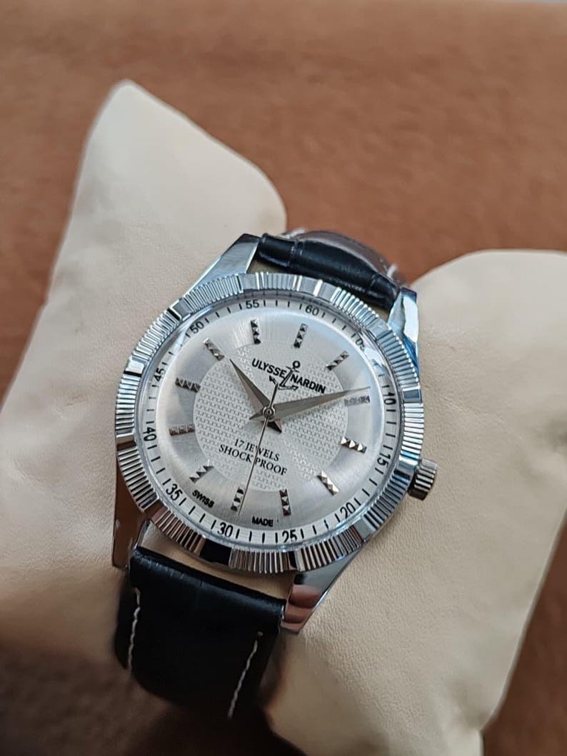 May include: Silver-toned Ulysse Nardin watch with a textured silver face, black leather band, and the text "17 JEWELS SHOCK PROOF SWISS MADE". The watch has a fluted bezel and is displayed on a cream-colored surface.