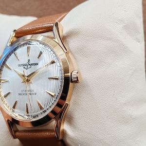 May include: A gold-tone wristwatch with a white dial featuring a vertical ribbed texture, gold-tone hands, and hour markers. The dial displays the text ULYSSE NARDIN, 17 JEWELS, SHOCK PROOF, and SWISS MADE. It includes a tan leather strap.