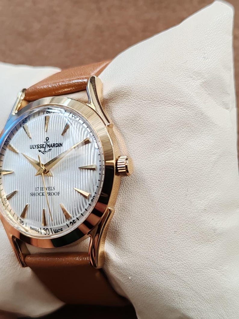 May include: A gold-tone wristwatch with a white dial featuring a vertical ribbed texture, gold-tone hands, and hour markers. The dial displays the text ULYSSE NARDIN, 17 JEWELS, SHOCK PROOF, and SWISS MADE. It includes a tan leather strap.