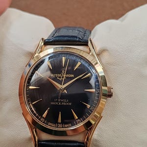 Puede incluir: Un reloj de pulsera dorado con esfera negra, manecillas e &iacute;ndices dorados. La esfera muestra el texto ULYSSE NARDIN, 17 JEWELS, SHOCK PROOF y SWISS MADE. Incluye una correa de cuero negro con un acabado texturizado.