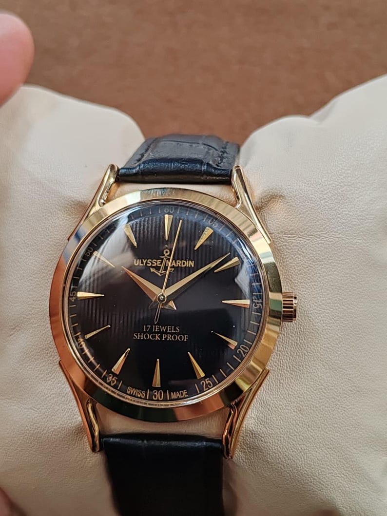 Puede incluir: Un reloj de pulsera dorado con esfera negra, manecillas e &iacute;ndices dorados. La esfera muestra el texto ULYSSE NARDIN, 17 JEWELS, SHOCK PROOF y SWISS MADE. Incluye una correa de cuero negro con un acabado texturizado.