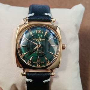 K&ouml;nnte beinhalten: Eine goldfarbene Armbanduhr mit quadratischem Geh&auml;use und gr&uuml;nem Sonnenschliff-Zifferblatt mit goldfarbenen Indizes und Zeigern. Das Zifferblatt zeigt den Text ULYSSE NARDIN, 17 JEWELS, SHOCK PROOF und SWISS MADE. Schwarzes Lederarmband mit wei&szlig;en N&auml;hten.