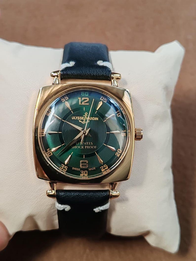 K&ouml;nnte beinhalten: Eine goldfarbene Armbanduhr mit quadratischem Geh&auml;use und gr&uuml;nem Sonnenschliff-Zifferblatt mit goldfarbenen Indizes und Zeigern. Das Zifferblatt zeigt den Text ULYSSE NARDIN, 17 JEWELS, SHOCK PROOF und SWISS MADE. Schwarzes Lederarmband mit wei&szlig;en N&auml;hten.