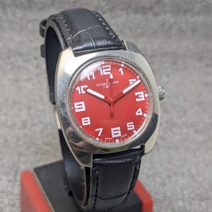 May include: A wristwatch with a bright red dial, white Arabic numerals, and white hands. The watch features a silver-toned cushion-shaped case and a black leather strap with a textured finish. The dial displays the Ulysse Nardin logo and text.