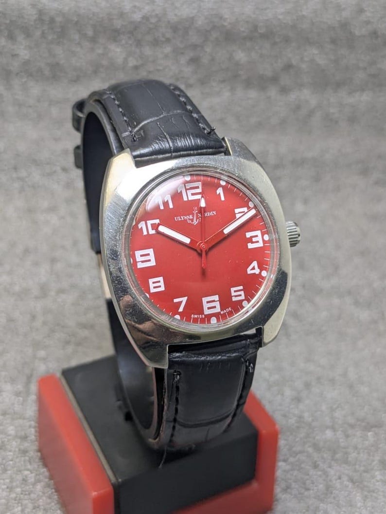 May include: A wristwatch with a bright red dial, white Arabic numerals, and white hands. The watch features a silver-toned cushion-shaped case and a black leather strap with a textured finish. The dial displays the Ulysse Nardin logo and text.