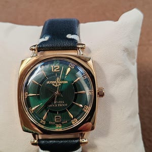 K&ouml;nnte beinhalten: Eine goldfarbene Armbanduhr mit quadratischem Geh&auml;use und dunkelgr&uuml;nem Zifferblatt mit Sonnenschliff. Das Zifferblatt zeigt goldene Indizes, arabische Ziffern bei 12 und 6 sowie den Schriftzug Ulysse Nardin, 17 Jewels, Shock Proof und Swiss Made.