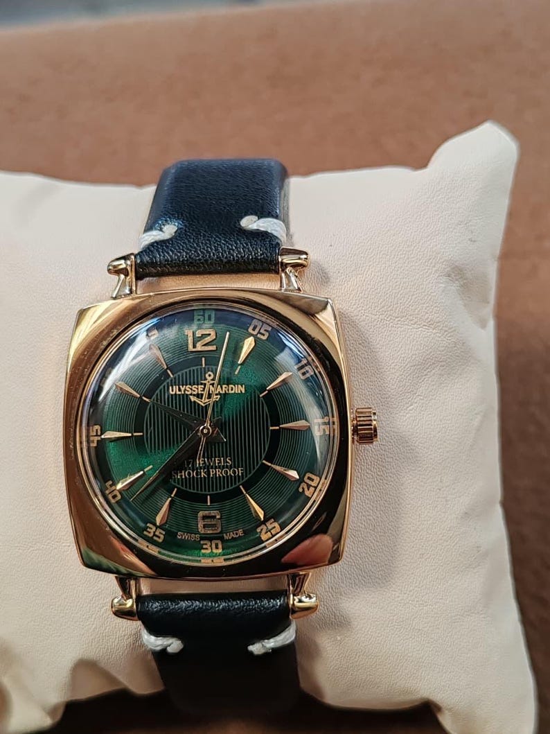 K&ouml;nnte beinhalten: Eine goldfarbene Armbanduhr mit quadratischem Geh&auml;use und dunkelgr&uuml;nem Zifferblatt mit Sonnenschliff. Das Zifferblatt zeigt goldene Indizes, arabische Ziffern bei 12 und 6 sowie den Schriftzug Ulysse Nardin, 17 Jewels, Shock Proof und Swiss Made.