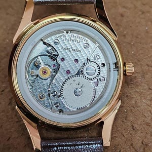 May include: A gold-tone wristwatch with an exposed mechanical movement visible through the case back. The movement is engraved with "SWISS SEVENTEEN 17 JEWELS" and "ULYSSE NARDIN" over a textured pattern. It features a brown leather strap.