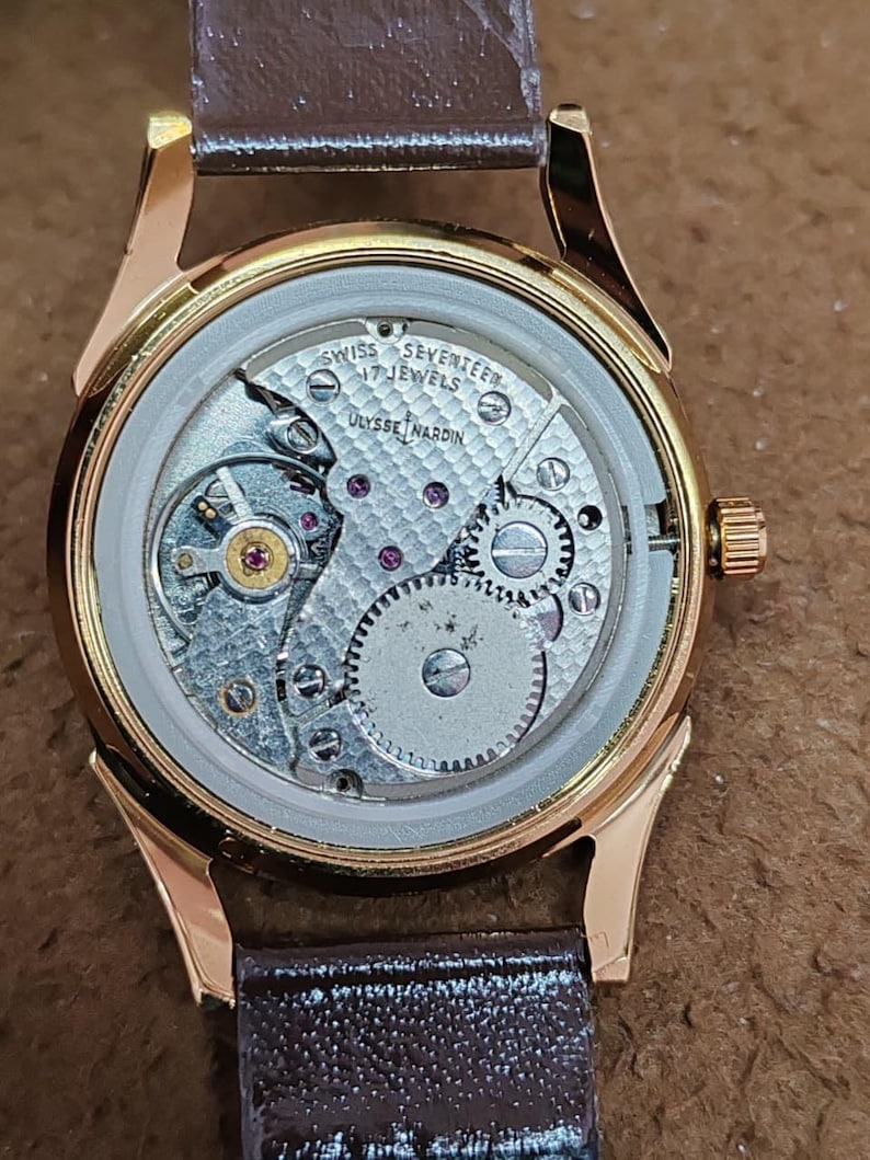 May include: A gold-tone wristwatch with an exposed mechanical movement visible through the case back. The movement is engraved with "SWISS SEVENTEEN 17 JEWELS" and "ULYSSE NARDIN" over a textured pattern. It features a brown leather strap.