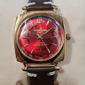 May include: A gold-tone wristwatch with a square case and a deep red sunburst dial. The dial features gold-tone indices, the text Ulysse Nardin, 17 Jewels, Shock Proof, and Swiss Made. It has a brown leather strap with white stitching.