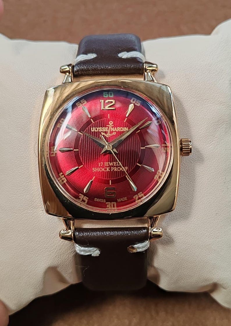 May include: A gold-tone wristwatch with a square case and a deep red sunburst dial. The dial features gold-tone indices, the text Ulysse Nardin, 17 Jewels, Shock Proof, and Swiss Made. It has a brown leather strap with white stitching.