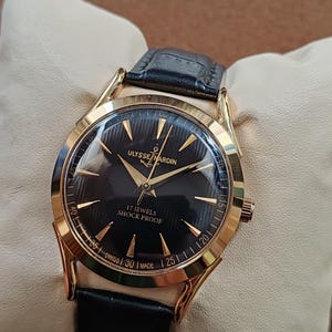 Puede incluir: Un reloj de pulsera dorado con esfera negra, manecillas doradas e &iacute;ndices dorados. La esfera muestra el texto ULYSSE NARDIN, 17 JEWELS, SHOCK PROOF y SWISS MADE. Tiene una correa de cuero negro con acabado texturizado y una caja redonda dorada.