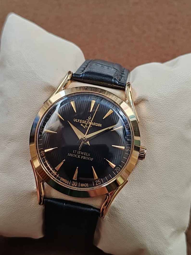 Puede incluir: Un reloj de pulsera dorado con esfera negra, manecillas doradas e &iacute;ndices dorados. La esfera muestra el texto ULYSSE NARDIN, 17 JEWELS, SHOCK PROOF y SWISS MADE. Tiene una correa de cuero negro con acabado texturizado y una caja redonda dorada.