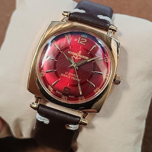 May include: A gold-tone wristwatch with a square case and a vibrant red sunburst dial. The dial features gold indices, the Ulysse Nardin logo with an anchor, and the text 17 Jewels Shock Proof. It has a dark brown leather strap with white stitching.