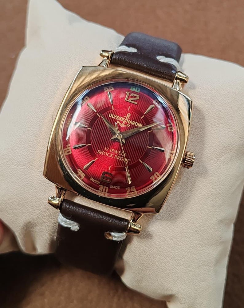 May include: A gold-tone wristwatch with a square case and a vibrant red sunburst dial. The dial features gold indices, the Ulysse Nardin logo with an anchor, and the text 17 Jewels Shock Proof. It has a dark brown leather strap with white stitching.