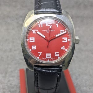 May include: A wristwatch with a silver-toned cushion-shaped case and a vibrant red dial. The face features white Arabic numerals, white hands, and the text Ulysse Nardin with an anchor logo. It includes a black leather strap with a textured finish.