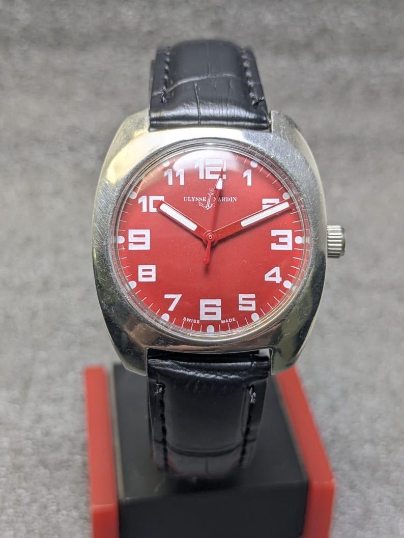 May include: A wristwatch with a silver-toned cushion-shaped case and a vibrant red dial. The face features white Arabic numerals, white hands, and the text Ulysse Nardin with an anchor logo. It includes a black leather strap with a textured finish.