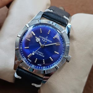 May include: A Ulysse Nardin wristwatch with a deep blue dial, silver-tone hands, and silver hour markers. The watch features a fluted silver bezel, a black leather strap with white stitching, and text reading Ulysse Nardin, Jewels, Shock Proof, Swiss Made.