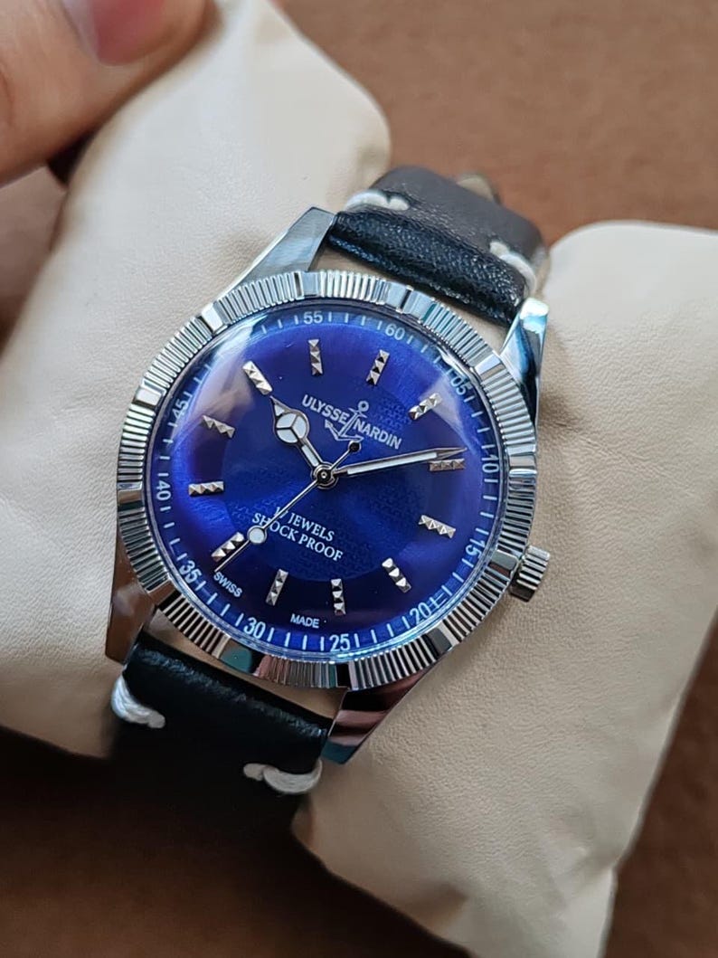 May include: A Ulysse Nardin wristwatch with a deep blue dial, silver-tone hands, and silver hour markers. The watch features a fluted silver bezel, a black leather strap with white stitching, and text reading Ulysse Nardin, Jewels, Shock Proof, Swiss Made.