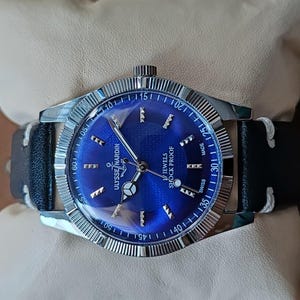 May include: A silver-tone Ulysse Nardin wristwatch with a vibrant blue sunburst dial, silver hour markers, and a black leather strap with white stitching. The watch face features the text Ulysse Nardin, 17 Jewels, Shock Proof, and Swiss Made.