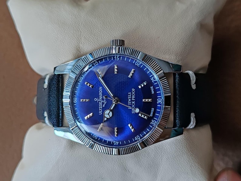 May include: A silver-tone Ulysse Nardin wristwatch with a vibrant blue sunburst dial, silver hour markers, and a black leather strap with white stitching. The watch face features the text Ulysse Nardin, 17 Jewels, Shock Proof, and Swiss Made.