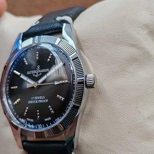 K&ouml;nnte beinhalten: Eine silberfarbene Armbanduhr mit schwarzem Zifferblatt, das das Ulysse Nardin-Logo mit einem Anker sowie silberne Zeiger und Indizes zeigt. Auf dem Zifferblatt steht 17 JEWELS SHOCK PROOF und SWISS MADE. Sie hat eine geriffelte L&uuml;nette und ein schwarzes Lederarmband.