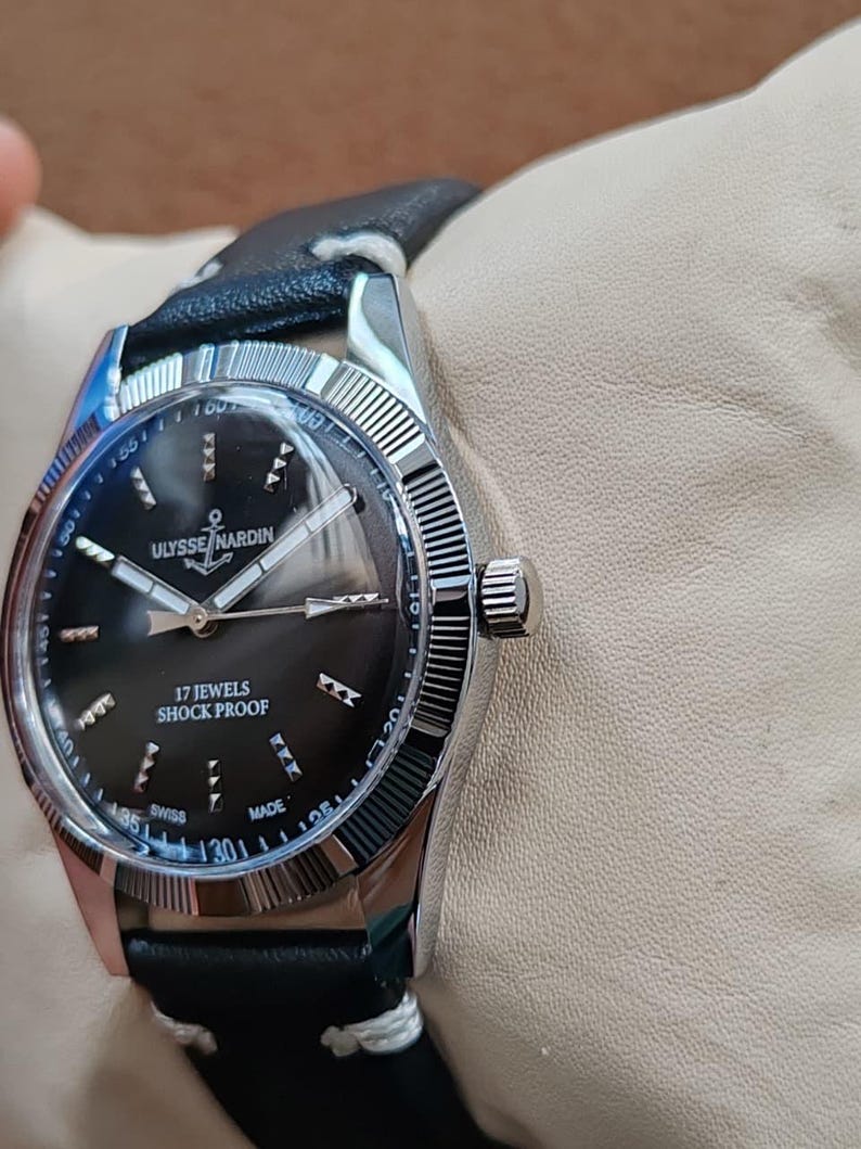 K&ouml;nnte beinhalten: Eine silberfarbene Armbanduhr mit schwarzem Zifferblatt, das das Ulysse Nardin-Logo mit einem Anker sowie silberne Zeiger und Indizes zeigt. Auf dem Zifferblatt steht 17 JEWELS SHOCK PROOF und SWISS MADE. Sie hat eine geriffelte L&uuml;nette und ein schwarzes Lederarmband.