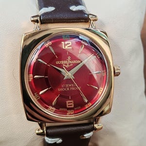May include: A gold-tone wristwatch with a square case and a deep red sunburst dial. The dial features gold-tone indices, the text ULYSSE NARDIN with an anchor logo, 17 JEWELS, SHOCK PROOF, and SWISS MADE. It has a dark brown leather strap with white stitching.