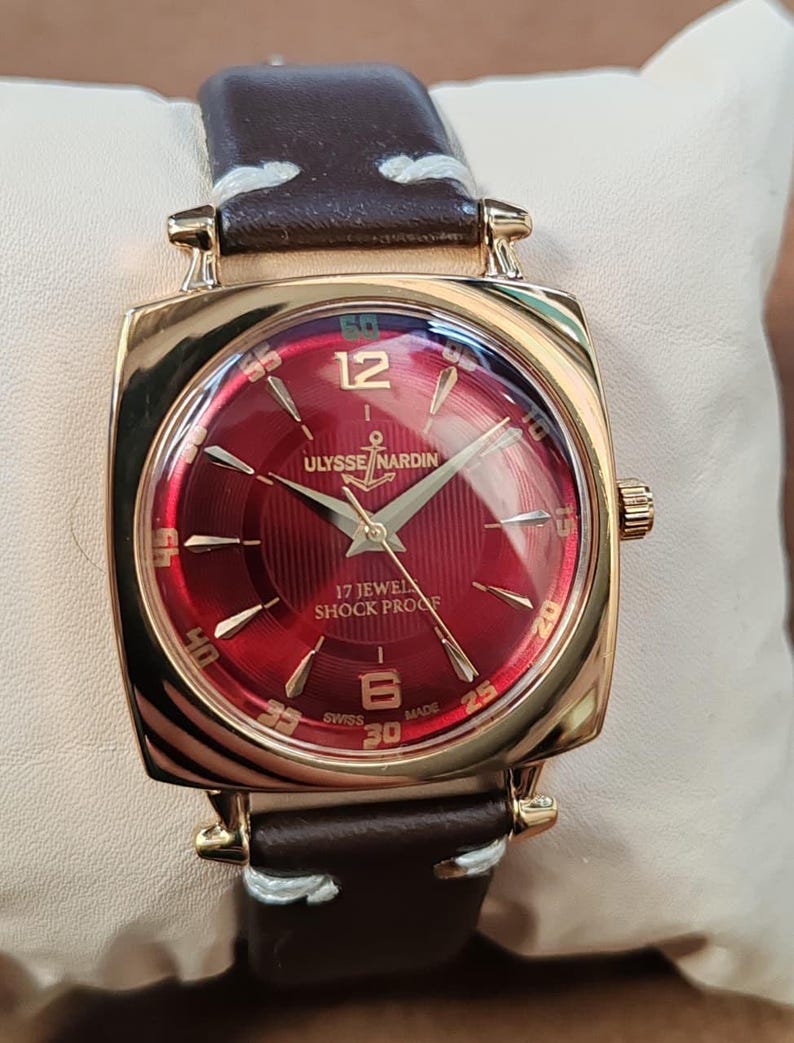 May include: A gold-tone wristwatch with a square case and a deep red sunburst dial. The dial features gold-tone indices, the text ULYSSE NARDIN with an anchor logo, 17 JEWELS, SHOCK PROOF, and SWISS MADE. It has a dark brown leather strap with white stitching.