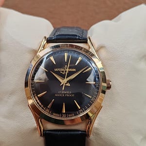 Puede incluir: Un reloj de pulsera dorado con esfera negra, manecillas doradas e &iacute;ndices dorados. La esfera muestra el texto ULYSSE NARDIN, un logotipo de ancla, 17 JEWELS, SHOCK PROOF y SWISS MADE. Cuenta con una correa de cuero negro con acabado texturizado.