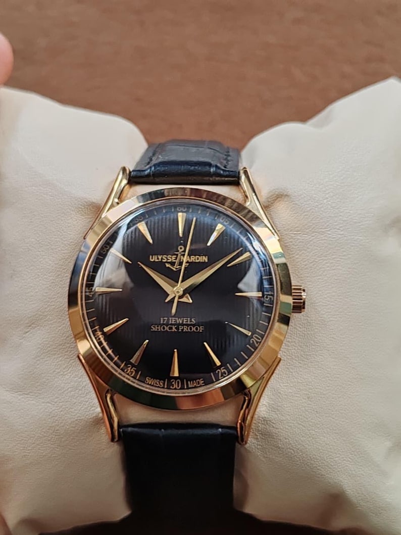 Puede incluir: Un reloj de pulsera dorado con esfera negra, manecillas doradas e &iacute;ndices dorados. La esfera muestra el texto ULYSSE NARDIN, un logotipo de ancla, 17 JEWELS, SHOCK PROOF y SWISS MADE. Cuenta con una correa de cuero negro con acabado texturizado.