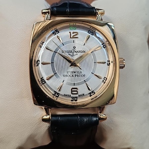 May include: A gold-tone wristwatch with a square case and a black leather strap. The silver dial features a vertical striped pattern, gold-tone hands, Arabic numerals at 12 and 6, and the text Ulysse Nardin, 17 Jewels, Shock Proof, and Swiss Made.
