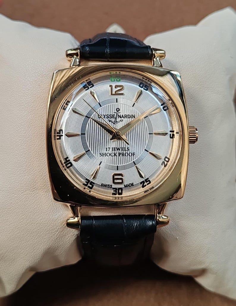 May include: A gold-tone wristwatch with a square case and a black leather strap. The silver dial features a vertical striped pattern, gold-tone hands, Arabic numerals at 12 and 6, and the text Ulysse Nardin, 17 Jewels, Shock Proof, and Swiss Made.
