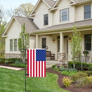 May include: A rectangular American flag with red and white stripes and a blue field of white stars. The flag is displayed on a black metal stand in a front yard with a house in the background.