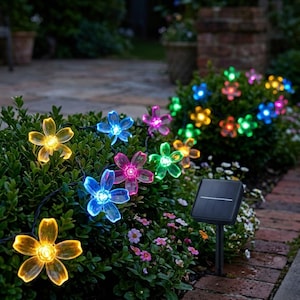 May include: A string of solar-powered flower lights in various colors, including yellow, blue, pink, and green, illuminates a garden. The lights are strung along a dark wire and are placed in a green bush. A small solar panel is visible.