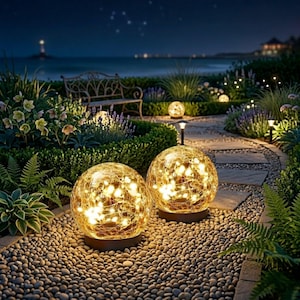 May include: Two illuminated glass globe lights with warm white fairy lights sit on a pebble path in a garden. The lights are surrounded by lush greenery and flowers. The scene is set at dusk with a view of the ocean and a lighthouse in the distance.