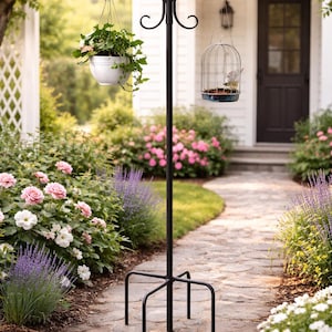 May include: A black metal bird feeder and plant stand with a hanging bird cage and a white planter filled with greenery. The stand is on a stone path surrounded by flowers and bushes.