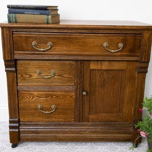 May include: A medium-brown wooden cabinet featuring one wide top drawer, two smaller stacked drawers on the left, and a single door on the right. Each drawer has two ornate brass-toned metal handles, and the door has a small round knob.