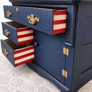 May include: A navy blue painted wooden chest with three open drawers and one cabinet door. The drawer sides feature horizontal red and white stripes. The chest includes ornate gold-toned metal drawer pulls and brass-colored hinges on the door.