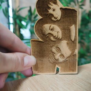 May include: A wooden plaque featuring three engraved faces. The faces are arranged in a vertical stack, with the top face partially overlapping the middle face. The wood has a warm, natural tone.