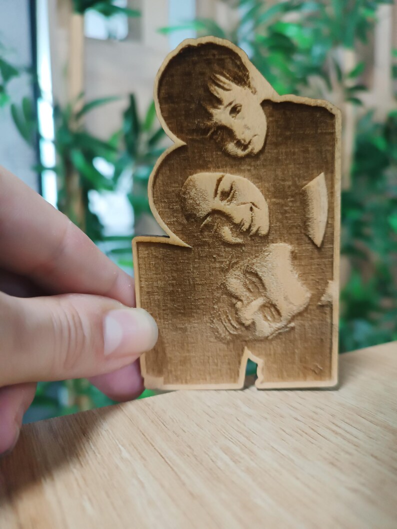 May include: A wooden plaque featuring three engraved faces. The faces are arranged in a vertical stack, with the top face partially overlapping the middle face. The wood has a warm, natural tone.