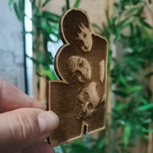 May include: A wooden cutout featuring three engraved faces. The faces are of different sizes and appear to be portraits. The wood has a warm, brown tone, and the details of the faces are visible.