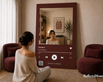 以下が含まれることがあります： バーガンディ色のフレームが付いた、音楽プレーヤーのインターフェースのようにデザインされた大きな長方形の鏡。鏡には、人、家具、アートワークのある部屋が映っています。「Perfect」と「Ed Sheeran」の文字が見えます。