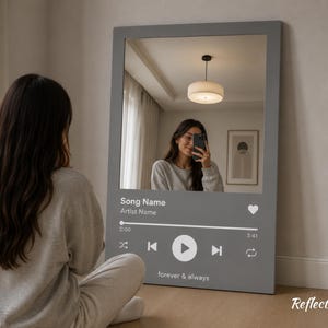 May include: A gray rectangular mirror with a music player interface design. The mirror reflects a person taking a selfie. The text on the mirror reads "Song Name", "Artist Name", "forever & always". The mirror is leaning against a wall.