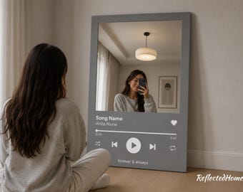 以下が含まれることがあります： 音楽プレーヤーのインターフェースデザインが施された灰色の長方形の鏡。鏡には自撮りをする人が映っています。鏡の文字は「Song Name」、「Artist Name」、「forever & always」です。鏡は壁に立てかけられています。