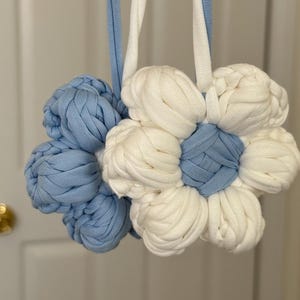 May include: Two flower-shaped ornaments crafted from braided fabric strips. One flower is light blue with a white center, and the other is white with a light blue center. Each features a long, matching fabric strap for hanging, made of woven material.