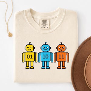 May include: A cream-colored t-shirt featuring a graphic of three cartoon robots in yellow, blue, and orange. Each robot displays a binary number on its chest: 01, 10, and 11. The shirt has a Comfort Colors brand label at the neck, size M.